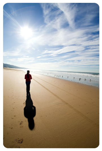 A woman walking alone along a wide sunlit beach, her long shadow stretching behind her.